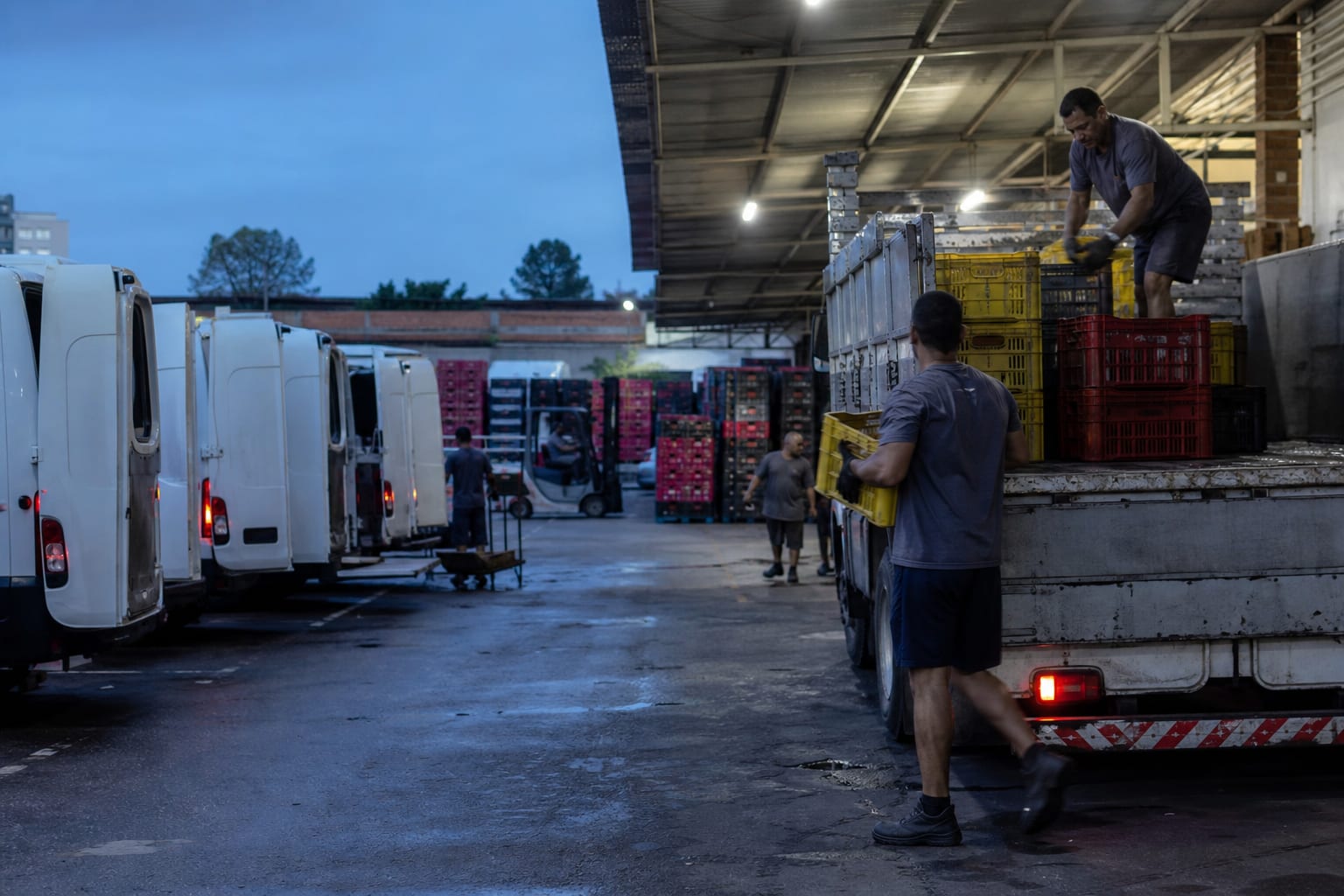 Vans brancas e caminhão leve sendo carregados com engradados no pátio de uma distribuidora de bebidas ao amanhecer