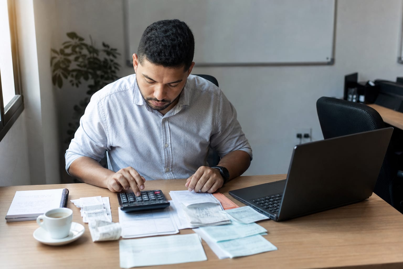 Gestor com calculadora, notebook e notas de oficina espalhadas sobre a mesa no fim da tarde