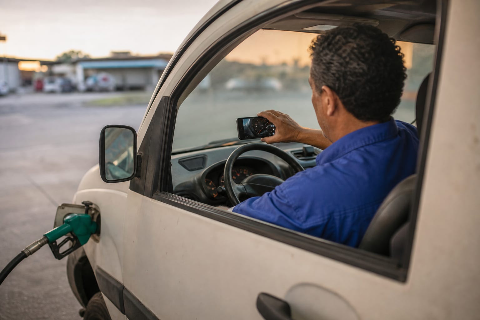 Motorista de uniforme azul fotografa com o celular o painel de instrumentos de uma van branca durante o abastecimento em um posto no fim da tarde